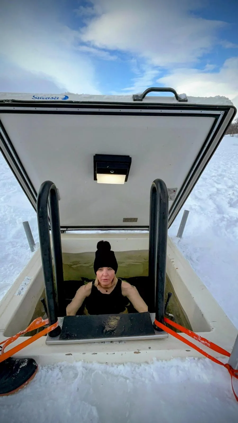 Sam sitting in an ice plunge pool cut into the snow, wearing a woolly hat